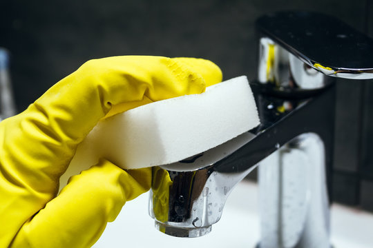 A hand in a yellow rubber glove wipes a dirty chrome metal faucet with a white melamine sponge. Cleaning in a modern bathroom. Selective focus. Closeup view