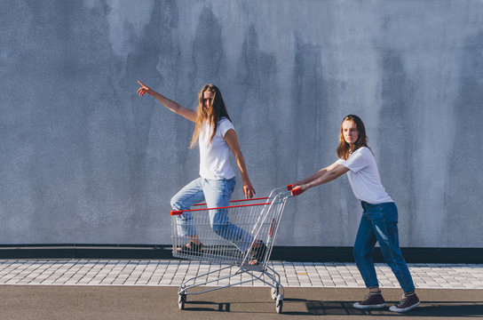 Two Happy Hipster Girls Staying On Shopping Cart Outdoors