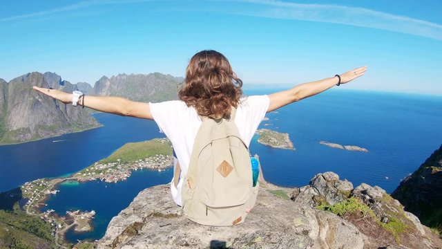 The young woman sitting at the edge of a cliff with a top view to the Reine, Lofoten Islands, Norway.