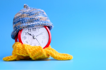 red round alarm clock in knitted wool blue hat and yellow scarf on a blue background. winter time...