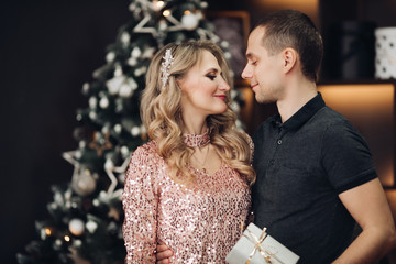 Stock photo of handsome couple in festive clothes celebrating New Year with glasses of wine or champagne. They are posing with drinks close to each other standing against Christmas interior. Golden