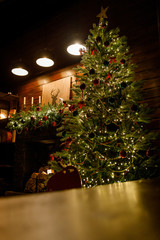 Christmas and New Year interior - blur background: fireplace, lamps, green Christmas tree, brown leather sofa, gifts, candles, moose rocking chair. Lots of lights glowing in the dark.