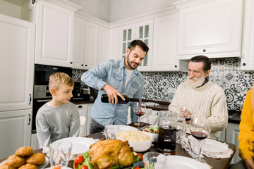 Young handsome man pouring wine into glass at served table while family having thanksgiving celebration at home