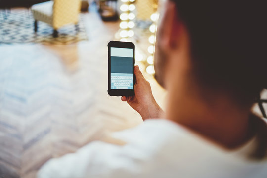 Back view of millennial male using mobile phone for chatting in social networks via app, cropped image of hipster guy holding smartphone while reading information from web page at home interior.