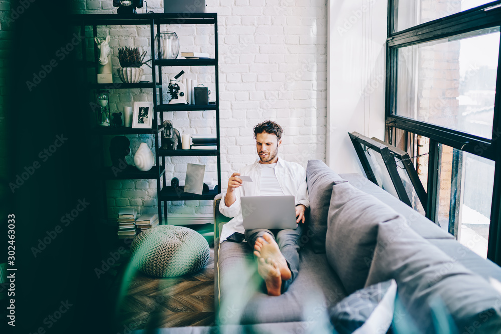 Poster cheerful hipster guy resting on sofa in modern designed living room making online shopping via lapto