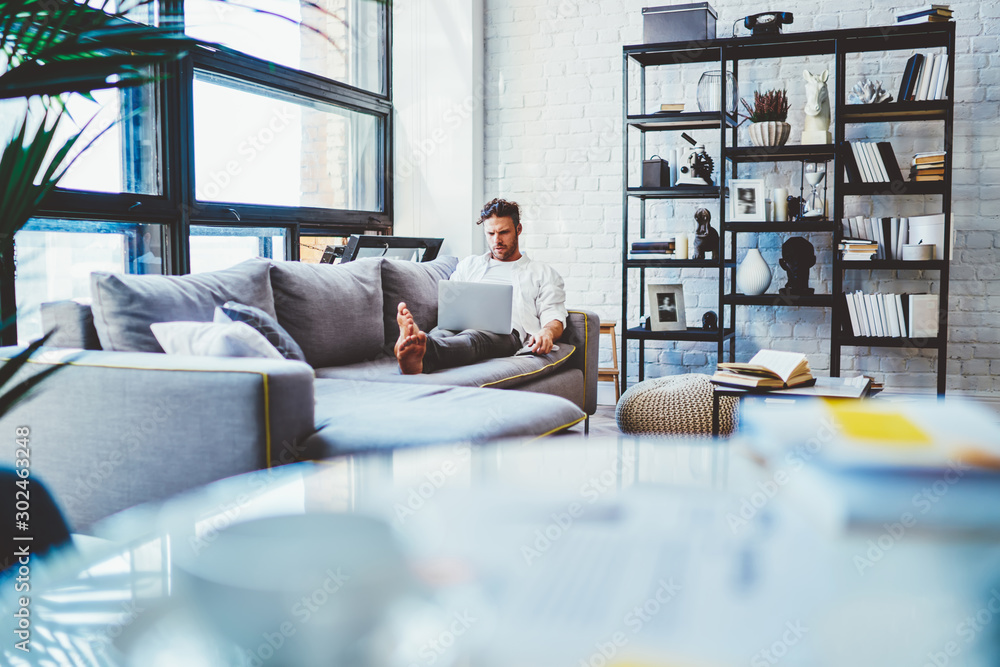 Poster millennial man lying on sofa with laptop computer spending time for messaging with followers from so
