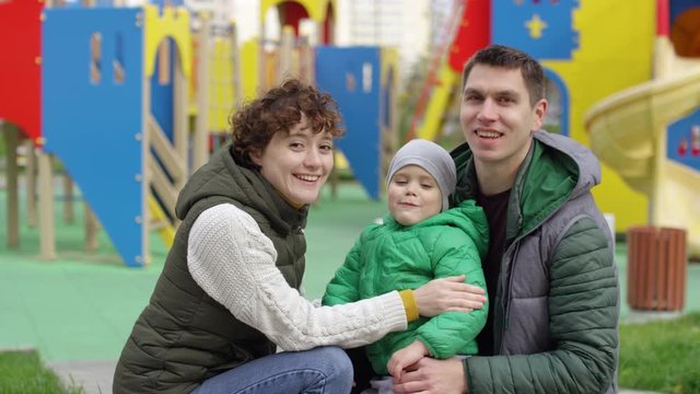Portrait shot of 30-something Caucasian husband, wife and 4-year-old son posing in front of urban playground, looking at camera, boy making funny face and puffing cheeks, and everybody laughing