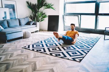 Young man sitting on carpet in modern apartment and doing morning exercises during yoga at home...