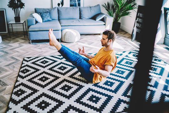 Experienced young man in sportive wear holding legs up sitting on carpet  during meditation at home interior.Hipster guy lead healthy lifestyle and  doing morning exercises in modern apartment Stock Photo | Adobe