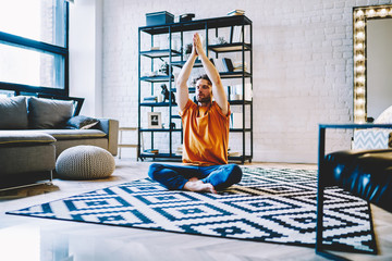 Calm young bearded man sitting in lotus pose on cozy carpet and holding hands in namaste above head...