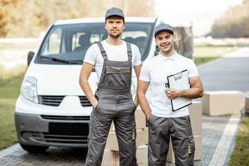 Portrait of a two delivery men in uniform standing together with check list and cardboard boxes...