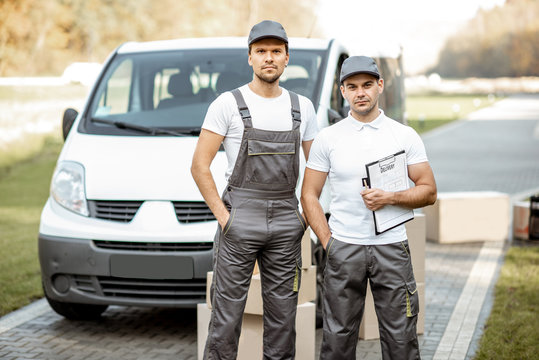 Portrait of a two delivery men in uniform standing together with check list and cardboard boxes near the cargo van vehicle outdoors