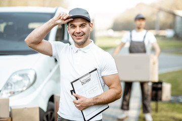 Portrait of delivery man in uniform standing with documents near the cargo van vehicle, colleague...