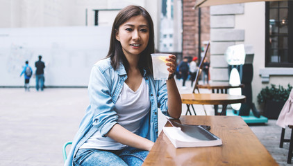 Portrait of positive Chinese female in casual denim shirt holding glass with cold lemon water and looking at camera during leisure day in downtown, attractive woman with beverage in hand outdoors