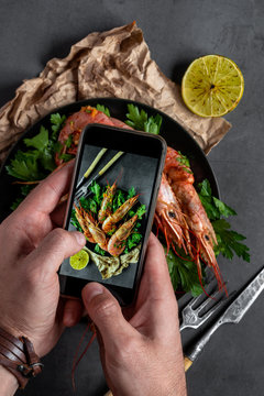 A man holds a smartphone in his hands and makes a photo of food. Tiger prawns on a gray background.