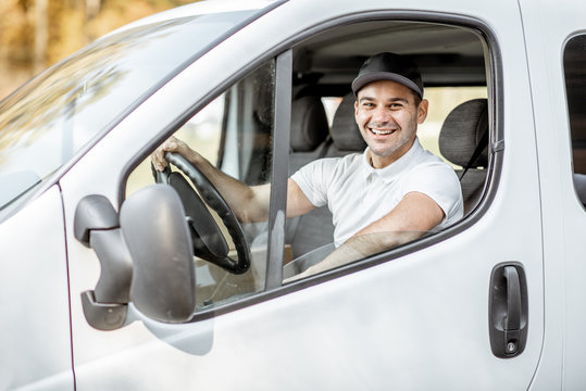 Portrait of a cheerful delivery driver in uniform looking out the window of the white cargo van vahicle, delivering goods by car