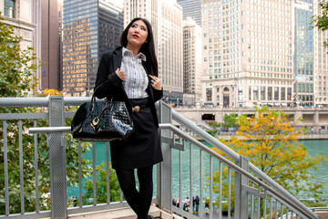 happy Asian business woman with long black hair wearing a trendy black blazer and skirt is walking confidently carrying a fashionable handbag while touring around the beautiful Chicago landscape 