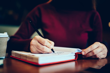 Close up of selective focus on woman's hands writing down information to red textbook using quality black pen.Cropped image of female sitting at wooden table and recording to notepad in coffee shop