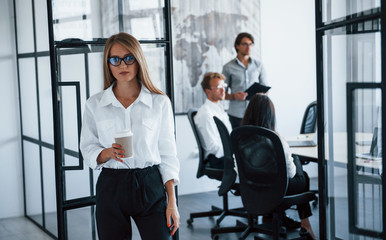Fototapeta na wymiar Brunette woman in glasses with cup of drink in hands stands in the office opposite employees