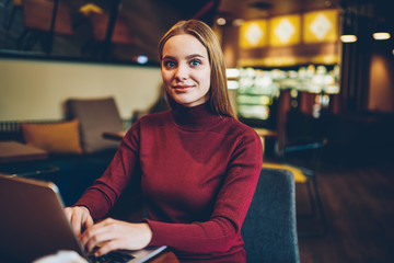 Fototapeta premium Portrait of positive skilled female student looking at camera during e learning with laptop computer in university cafeteria, young smiling woman freelancer working remotely in coworking space