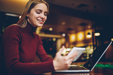 Happy hipster girl sharing media files via touch pad and laptop device using bluetooth connection indoors, smiling woman reading positive news in social networks via application on digital tablet