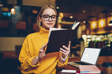 Attractive hipster girl reading interesting best seller sitting near laptop with mock up blank, beautiful female student in spectacles making research of information in textbook for education