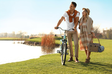 Young couple with bicycle and picnic basket near lake on sunny day