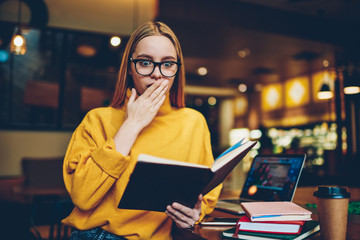 Young gorgeous emotional intelligent woman in glasses for eye protection impressed by interesting literature standing indoors.Good looking amazed hipster girl wondering while spending time in cafe
