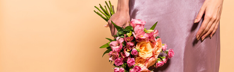 Fototapeta na wymiar partial view of elegant woman in violet satin dress holding bouquet of flowers isolated on beige, panoramic shot