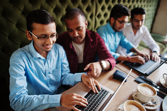 Group Of Four South Asian Men's Posed At Business Meeting In Cafe. Indians Work With Laptops Together Using Various Gadgets, Having Conversation.