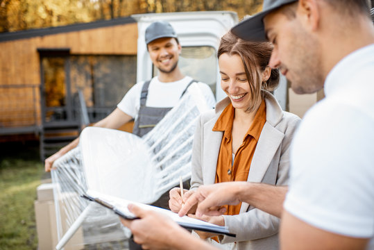 Delivery company employees unloading goods from a car trunk, delivering goods to a woman's home. Happy client signing delivery documents