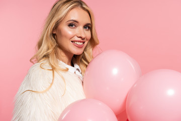 Obraz na płótnie Canvas smiling beautiful blonde woman in faux fur jacket holding pink balloons isolated on pink