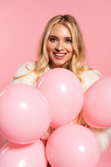 Fototapeta na wymiar happy beautiful blonde woman in faux fur jacket holding pink balloons isolated on pink
