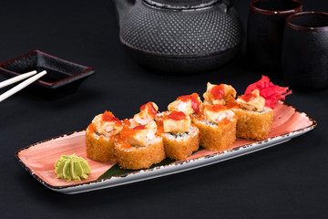 Close up of assorted baked sushi set with salmon, shrimp, caviar, mussels, tuna, cream cheese and hot sauce and chopsticks, soy on black background. Hot spicy rolls and green tea teapot