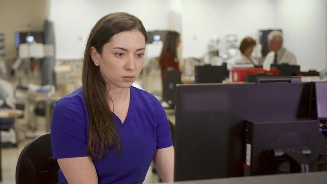 A young brunette nurse works on a computer in a hospital nurse's station. She is scheduling and reviewing patient information in her day to day tasks.