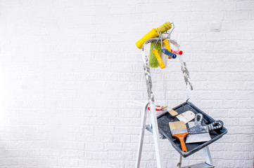 Painted metal stepladder with laid out on it rollers, brushes, colored spatulas, black tray and a bucket of paint.