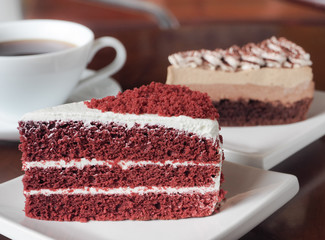 Slice of red velvet cake on a white plate. Close up of red velvet chocolate cake and tiramisu coffee cake with a cup of hot coffee on table.