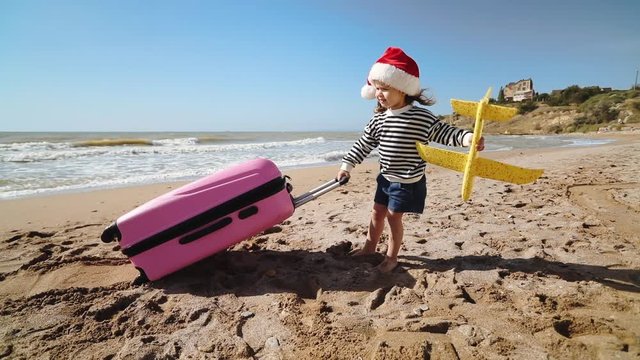 little girl wearing red Christmas hat holds yellow toy airplane and pulls large bright pink suitcase on sunny sand beach