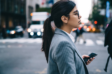 Side view of thoughtful business woman formally dressed walking to business meeting in financial district holding modern mobile cell gadget in hand, Latino female proud ceo with smartphone going