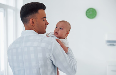 Stands and holds baby on the hands. Young pediatrician is in the clinic at daytime