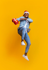 Fototapeta na wymiar Positive black guy in Santa hat jumping with Xmas present