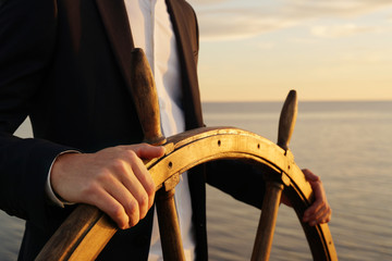 Businessman holding ship rudder and navigates in sunset light. - obrazy, fototapety, plakaty