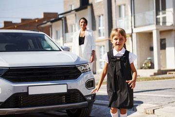 Mother with daughter in school uniform outdoors near white car