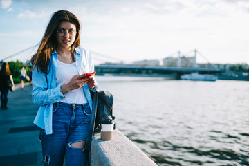 Attractive female tourist standing near river in city satisfied with spring weather while sightseeing, woman using mobile phone and 4G connection for blogging recreating walking near lake in town