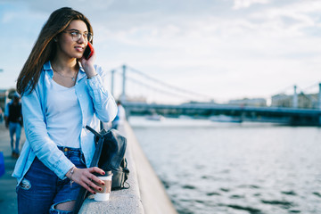 Young female tourist calling to friend in roaming while looking at beautiful river view on unrab setting, hipster girl having telephone conversation during free time outdoors enjoying spring