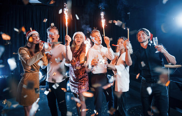 Fototapeta na wymiar Having fun with sparklers. Group of cheerful friends celebrating new year indoors with drinks in hands