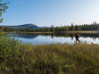 Fisherman man catching fish at lake Sjabatjakjaure in Beautiful sunny morning haze mist in Sweden Lapland nature. Mountains, birch trees, spruce forest. Blu sky