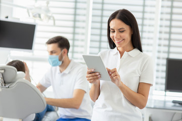 smiling female dentist using digital tablet while her collegue working with small parient on the background