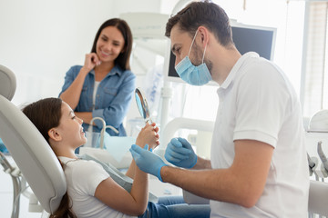 Fototapeta premium portrait of smiling girl looking in the mirror after dental procedure while mother standing near her for support