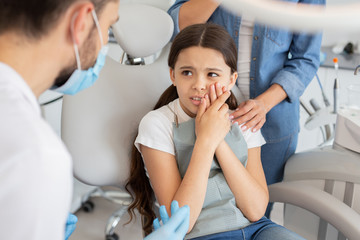 little patient with toothache and male doctor sitting in dental chair while mother standing near her for support